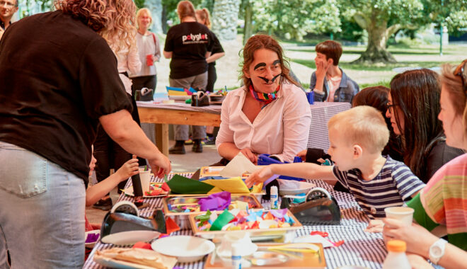 A Feast production photo. Two adults and a child gather around a table. There are tubs of coloured paper, pencils and decorations. Photo: Casey Horsfield.