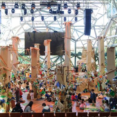 A Paper Planet Production Photo. Tall cardboard trees with cardboard branches dwarf a crowd of adults, performers and children who are decorating the forest area with paper and other decorations between the trees, making up a paper forest.