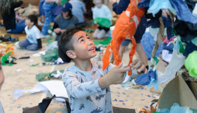 A Paper planet Production photo. A child wearing a long sleeved t-shirt holds a paper sculpture constructed from tissue paper and masking tape. In the background, children and adults lie on carpet surrounded by pieces of paper and tape. The child is in focus and the background is blurred.