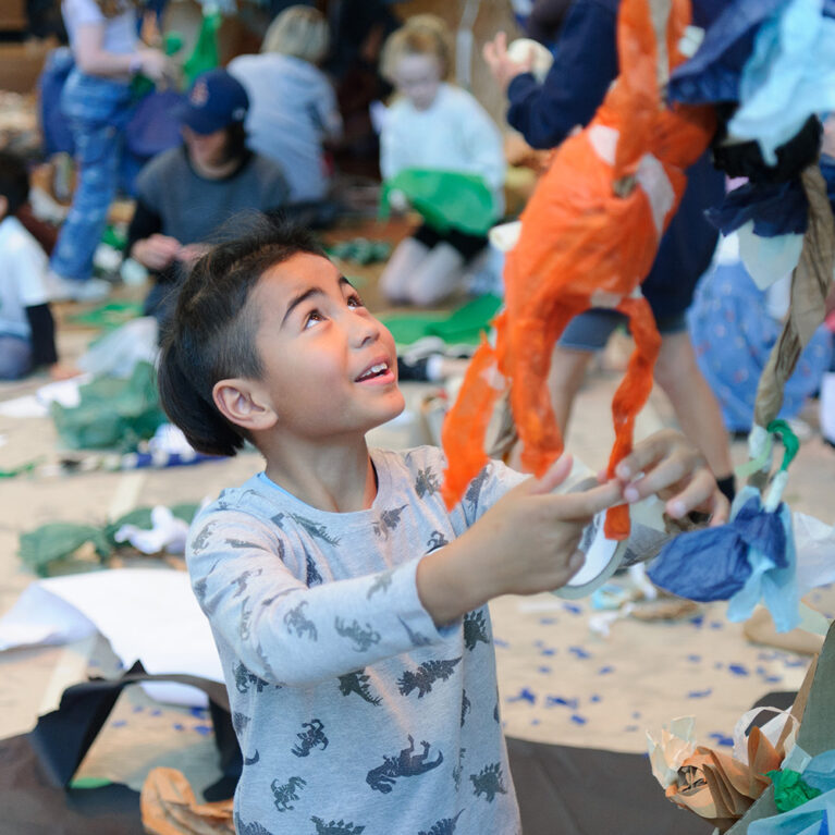 A Paper planet Production photo. A child wearing a long sleeved t-shirt holds a paper sculpture constructed from tissue paper and masking tape. In the background, children and adults lie on carpet surrounded by pieces of paper and tape. The child is in focus and the background is blurred.