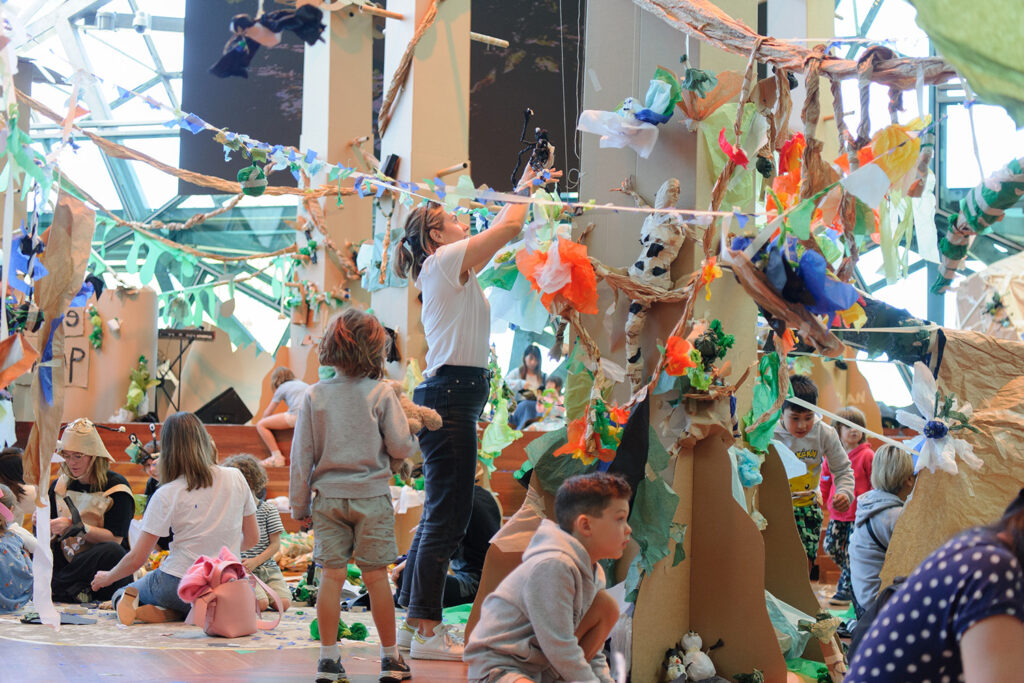 Paper Planet production photo: Sarah Walker. A forest of tall brown cardboard trees adorned with colourful tissue paper creatures and plants grows in a large space filled with natural light. Children and their families create and play with paper and tape among the trees.