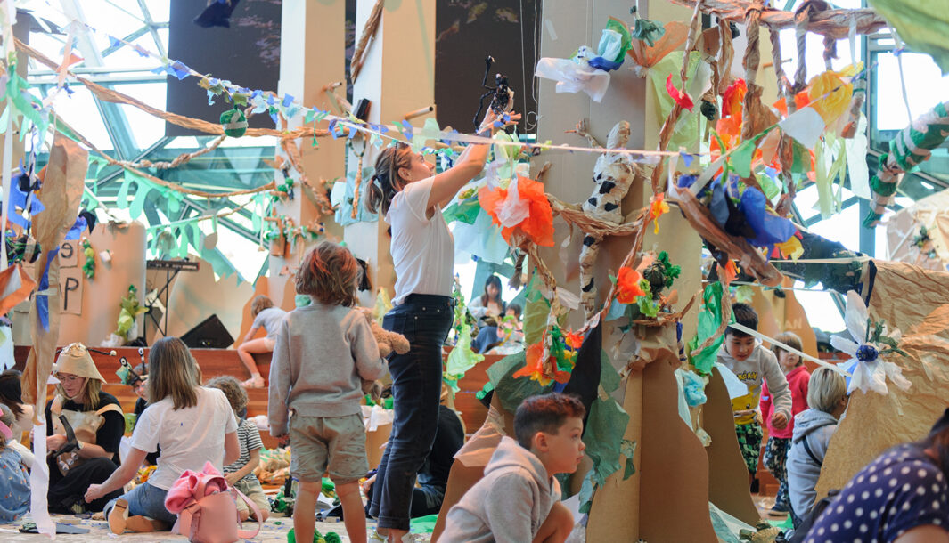 A Paper Planet production photo. A forest of tall brown cardboard trees adorned with colourful tissue paper creatures and plants grows in a large space filled with natural light. Children and their families create and play with paper and tape amongst the trees. Photographer: Sarah Walker