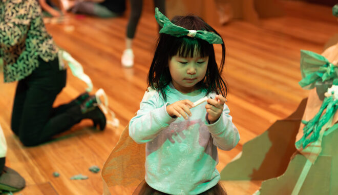 A Paper Planet Production photo. A child wears a paper head and plays with a piece of paper. They stand on a wood floor. Photo: Katje Ford.