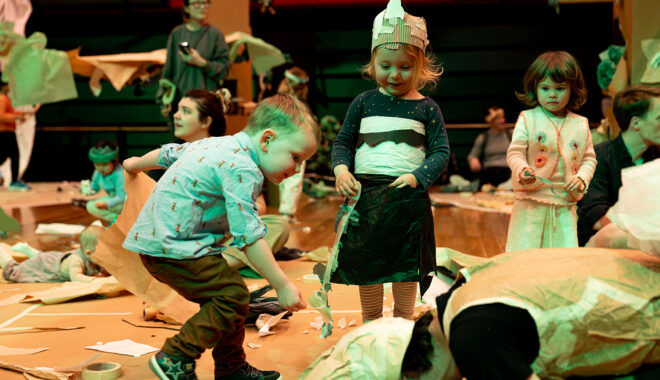A Paper Planet production photo. Two children play with a paper roll. The children stand on a wooden floor in a brightly-lit room. Photo: Katje Ford.