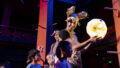 A Paper Planet production photo. A performer wearing rabbit ears made from paper holds an illuminated paper globe. A child looks up at them. Behind them is a cardboard paper tree that is partially illuminated by soft light. Photo: Katje Ford.