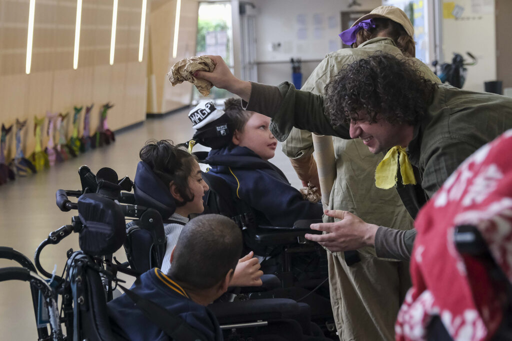 When the World Turns (schools) production photo: Suzanne Phoenix. Polyglot artists in khaki costumes engage with students in wheelchairs. They are in a school corridor.