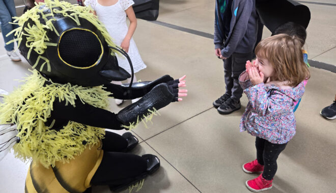 A Bees production photo. A performer in a bee costume outstretches their arms towards a child. The child covers their mouth in excitement. Photo: Libby Koba.