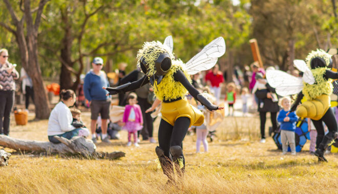 A Bees production photo. A performer dressed in a bee costume runs in a field of grass. They are being watched by children and families. Photo: Royal Botanic Gardens Victoria.