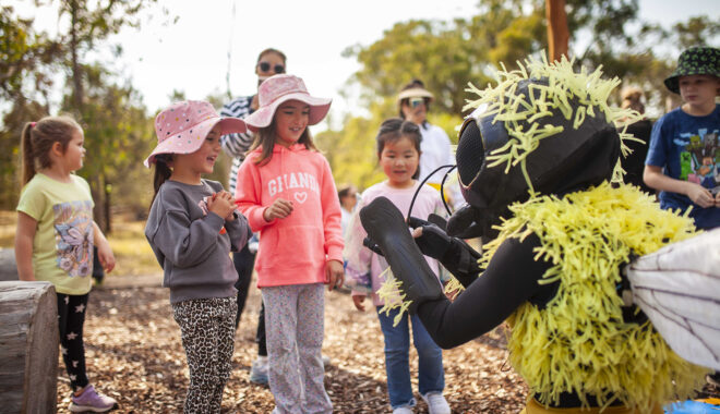 A Bees production photo. Three children stand across from a performer dresses as a bee, looking at the performer's hands. They stand on a field of mulch. Photo: Royal Botanic Gardens Victoria.