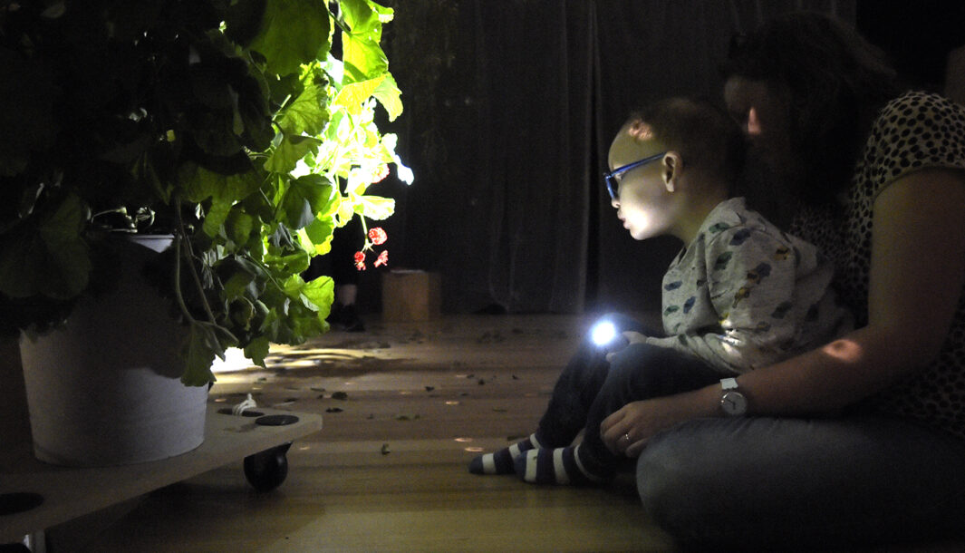A When the World Turns development photo. A child sits on their adult's lap on a wooden floor in a darkened space. They are pointing a small torch/flashlight at a large potted plant on a low trolley. Photographer: Carla Gottgens