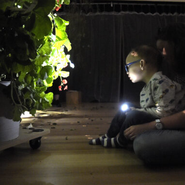 A When the World Turns development photo. A child sits on their adult's lap on a wooden floor in a darkened space. They are pointing a small torch/flashlight at a large potted plant on a low trolley. Photographer: Carla Gottgens