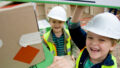 A We Built This City production photo. Two children wearing hi-vis vests and white hard hats smile at the camera while building with cardboard boxes.