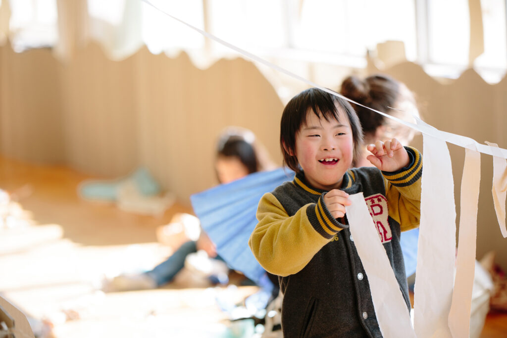 Paper Planet production photo: Ai Ueda. A child with dark hair, wearing a baseball jacket, joyfully hangs long strips of white paper on a suspended piece of tape. They are in a sunlit room lined with brown cardboard and other children creating with paper are visible in the background.
