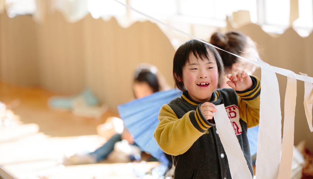 A Paper Planet production photo. A child with dark hair, wearing a baseball jacket, joyfully hangs long strips of white paper on a suspended piece of tape. They are in a sunlit room lined with brown cardboard and other children creating with paper are visible in the background.