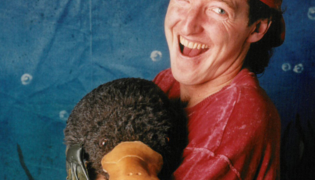 A 'Tadpole' production image. A man wearing a backwards red cap and a red shirt holds a puppet of a platypus against a blue background.