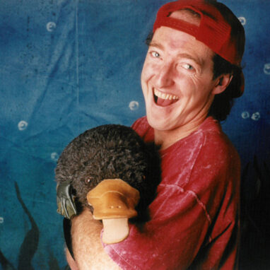 A 'Tadpole' production image. A man wearing a backwards red cap and a red shirt holds a puppet of a platypus against a blue background.