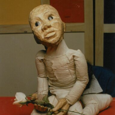 A 'Not The End of The World' production photo. A puppet made from wood and cloth with big, black pupils is seated and holds a rose with white petals in their hands.