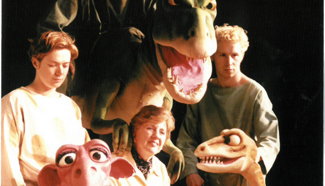 An 'Almost A Dinosaur' production photo. A woman poses between two dinosaur puppets. She is seated behind another large dinosaur puppet, which stands between two men. Behind the large dinosaur stands another man who is smiling.