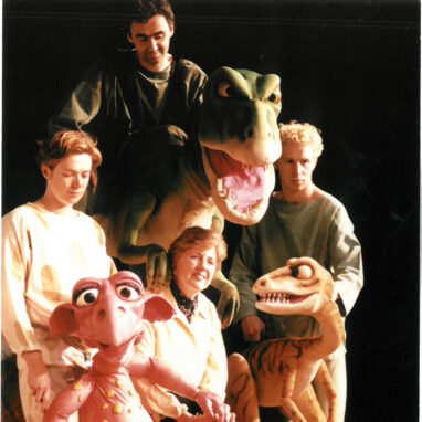 An 'Almost A Dinosaur' production photo. A woman poses between two dinosaur puppets. She is seated behind another large dinosaur puppet, which stands between two men. Behind the large dinosaur stands another man who is smiling.