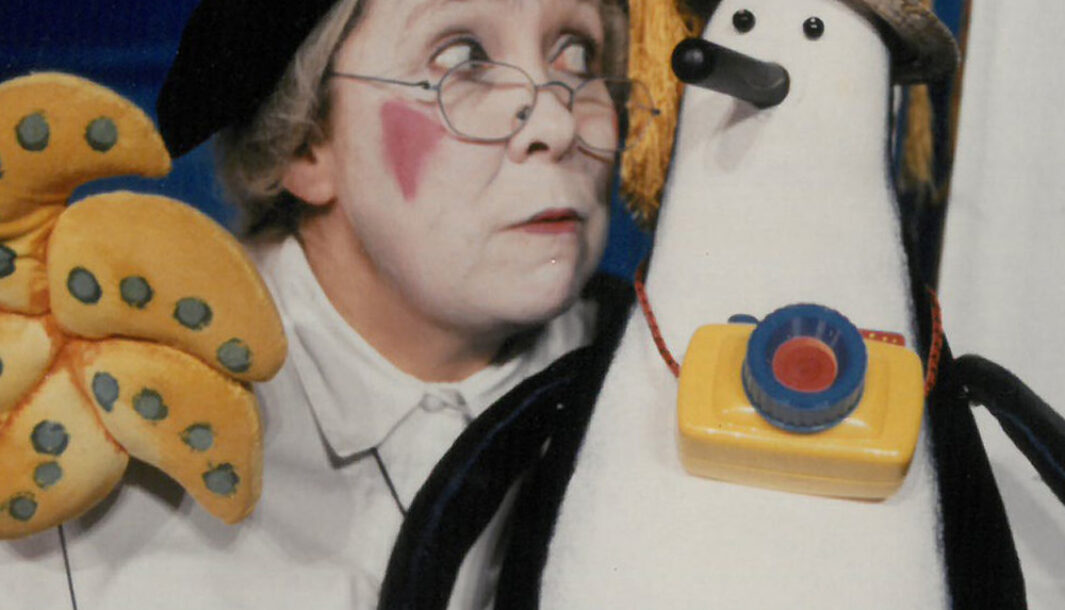 A Granny and the Sea Monster production photo. A person with white facial makeup and red cheeks holds a penguin shaped puppet. The puppet is wearing a yellow bag and a hat.