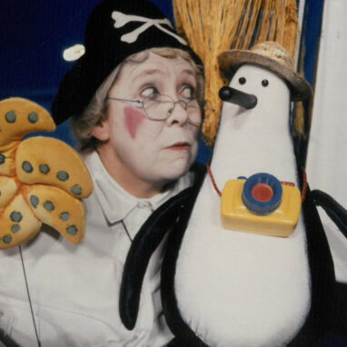 A Granny and the Sea Monster production photo. A person with white facial makeup and red cheeks holds a penguin shaped puppet. The puppet is wearing a yellow bag and a hat.