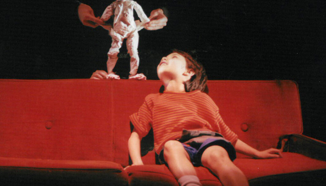 A 'Sunflowers' production photo. A child sits on a red couch and looks up at a puppet standing on the couch.