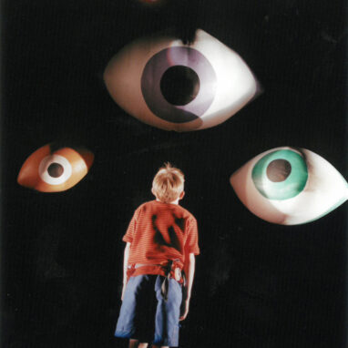 A Sunflowers Production photo. A child stands in front of a black wall. Three giant eyes hanging down in front of the wall look down at the child.