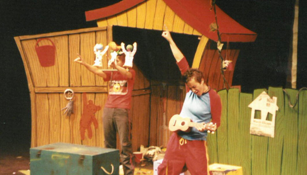 A Muckheap production photo. Two performers are performing on a stage in front of a wooden shed. The shed has a red roof and a green box placed in front of the shed. One performer is playing a ukelele and the other performer stands with puppets in the form of babies attached to a headpiece.