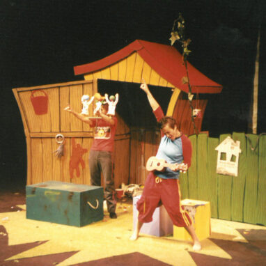 A Muckheap production photo. Two performers are performing on a stage in front of a wooden shed. The shed has a red roof and a green box placed in front of the shed. One performer is playing a ukelele and the other performer stands with puppets in the form of babies attached to a headpiece.