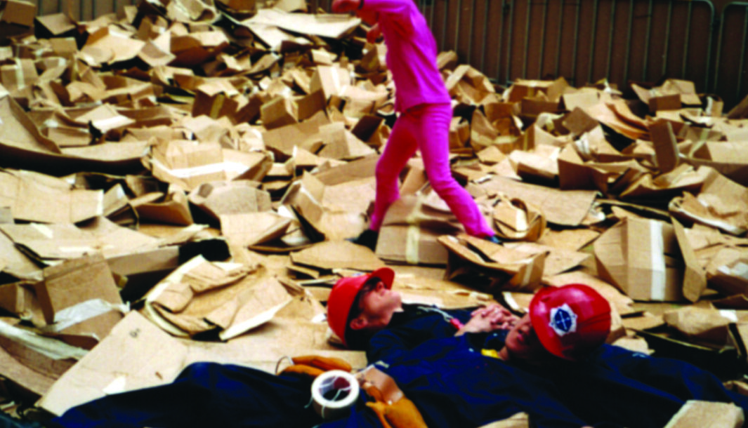 A We Built This City Production photo. A young person walks on a pile of cardboard boxes behind two performers. Performers lie on the cardboard boxes, and wear hard hats that cover their eyes.
