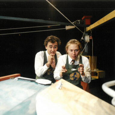 A 'The Mighty How' production photo. Two performers wearing white shirts and aprons look focused, crossing their fingers.