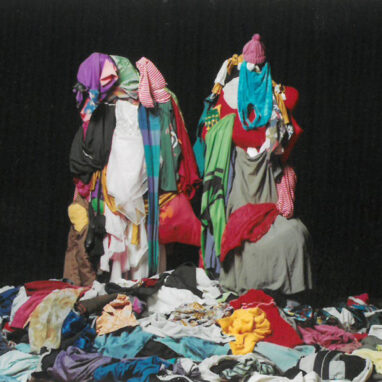 A Baggy Pants production photo. Two performers stand in front of a black background, covered in rags and clothes. They stand amongst a pile of clothes and rags.