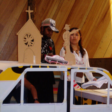 A Headhunter production photo. Two performers stand behind a makeshift car on stage. The car is decorated with warning tape. On top of the car are cardboard cutouts of the facade of a church, a car and a pointing man.