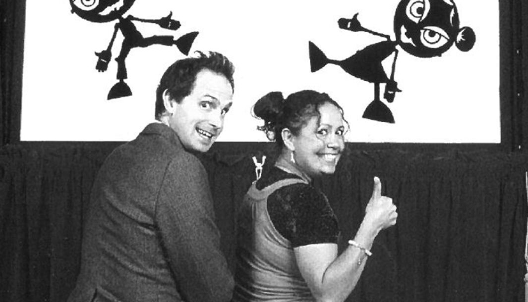 A Johnny Grimm production photo. Two performers smile and look behind their shoulders with their thumbs up. In front of the performers is an illuminated stage, where two shadow figures pose. Photo: Peter Marshall and Photo Montage by BAT Design.