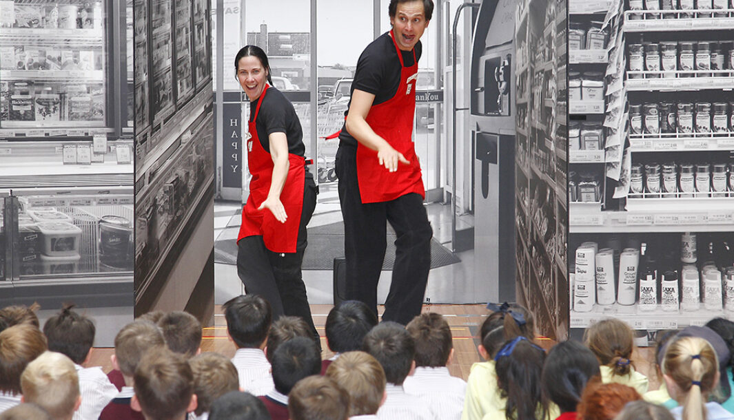 A Check Out! production photo. Two performers wear red aprons and pose sideways, facing an audience of children. Behind them are photo walls of shopping centres. Photo: Peter Marshall.