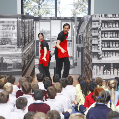 A Check Out! production photo. Two performers wear red aprons and pose sideways, facing an audience of children. Behind them are photo walls of shopping centres. Photo: Peter Marshall.