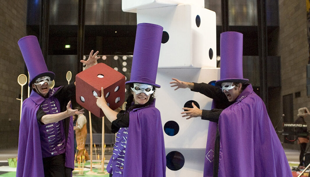 A 'The Big Game' production photo. Three performers stand in front of a pile of large dice blocks. They wear tall purple caps, purple capes and silver eye masks. The performer in the middle of the three holds a large red dice. Photographer: Gavin D Andrew.