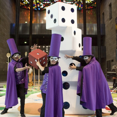 A 'The Big Game' production photo. Three performers stand in front of a pile of large dice blocks. They wear tall purple caps, purple capes and silver eye masks. The performer in the middle of the three holds a large red dice. Photographer: Gavin D Andrew.