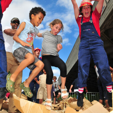 A 'We Built This City' production photo. Two children and a performer wearing blue overalls and a red t-shirt jump on cardboard boxes underneath a sunny sky.