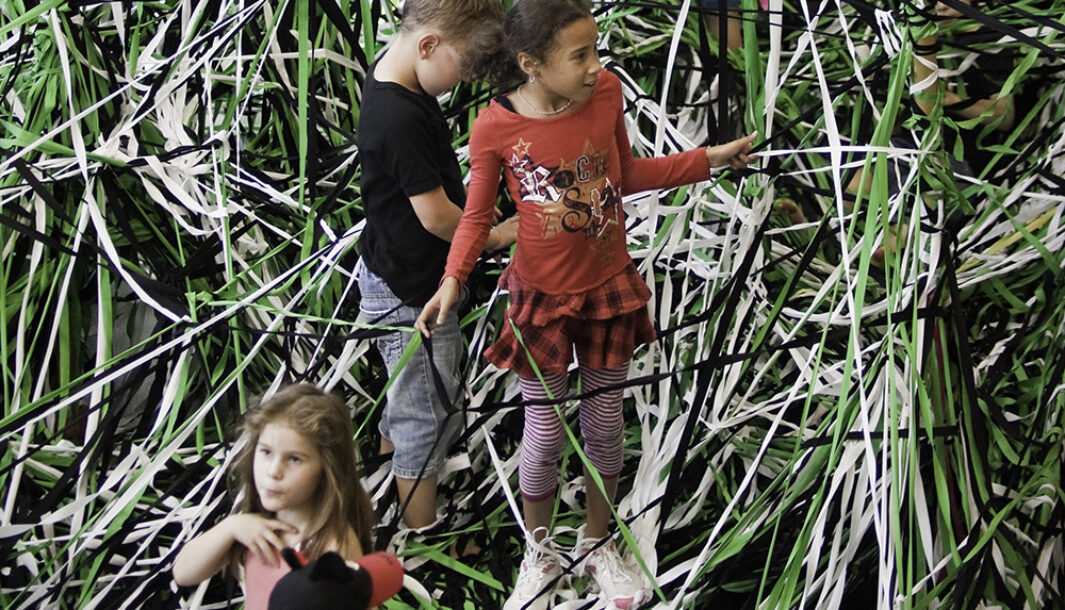 A Tangle production photo. Children stand on elastic string that is tangled in all directions. The string is coloured green, black and white. Photo; Aaron Walker.