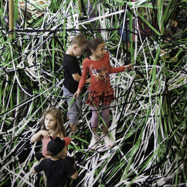 A Tangle production photo. Children stand on elastic string that is tangled in all directions. The string is coloured green, black and white. Photo; Aaron Walker.