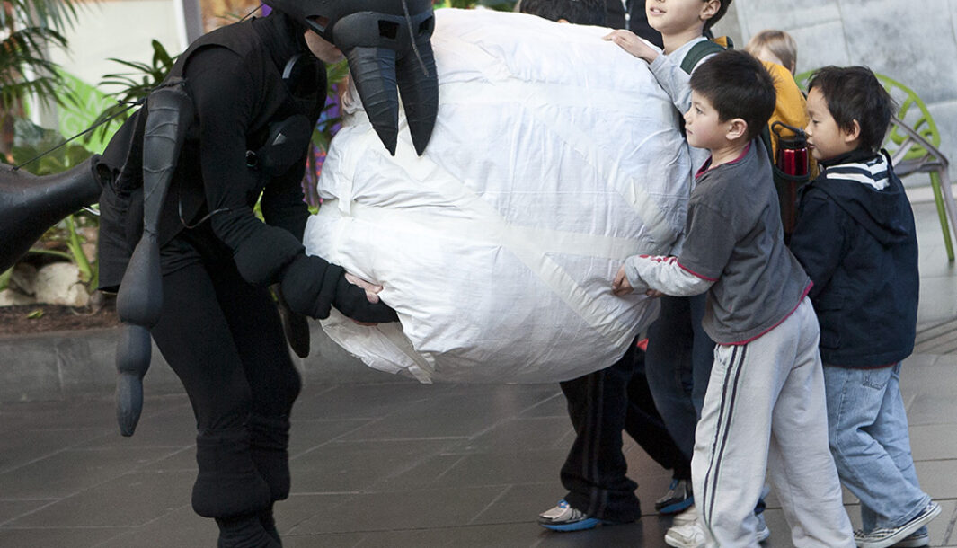 An Ants production photo. A performer dressed as an ant holds a large white crumb with other children who help to carry the crumb. Photo: Gavin D Andrew.