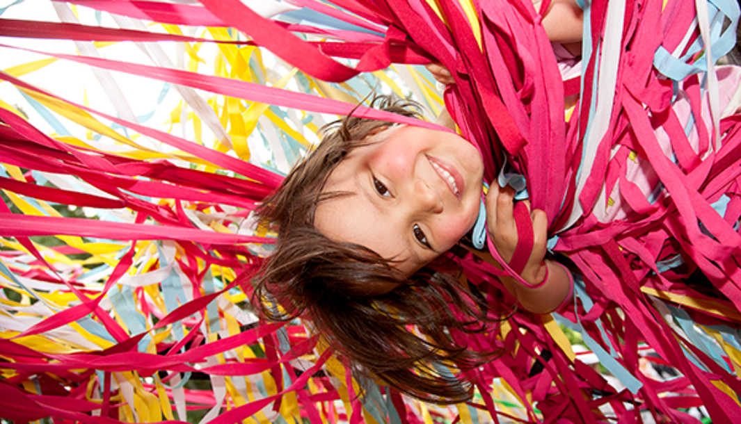 A Tangle production photo. A child with short dark hair is suspended upside down amongst hundreds of strands of colourful elastic. They are smiling at the camera.