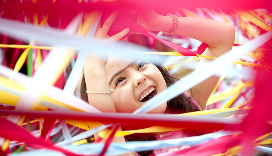 A Tangle production photo. A child is surrounded by hundreds of strands of colourful elastic. They are laughing and their hands are raised. Photographer: Wendy Kimpton
