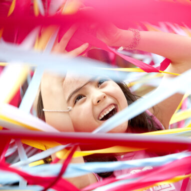 A Tangle production photo. A child is surrounded by hundreds of strands of colourful elastic. They are laughing and their hands are raised. Photographer: Wendy Kimpton