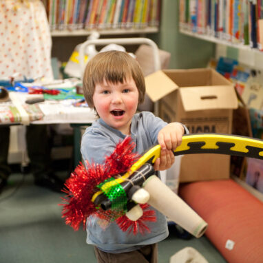A workshop photo. A small child holds up a creation made from assorted objects and materials, smiling widely at the camera.
