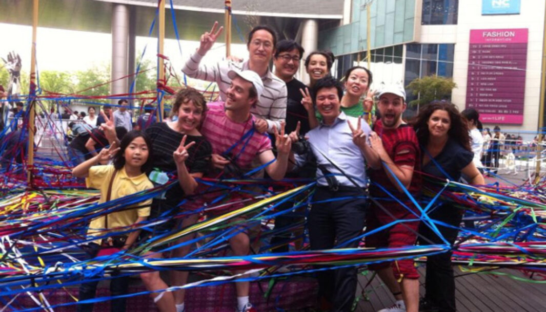 A Tangle Production photo. A group of adults and one child pose holding up the peace sign, surrounded by hundreds of weaved elastic strings wrapped around tall steel poles.