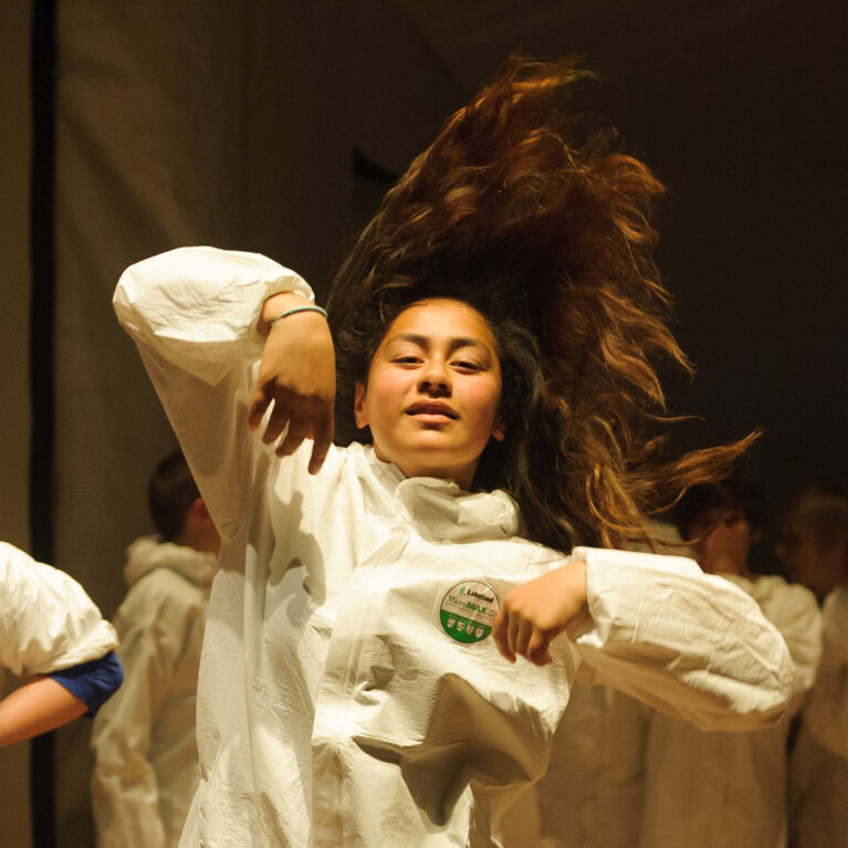 A Mahogany Rise school residency photo. A student wearing a white boiler suit is dancing, their long hair fanning out behind them.