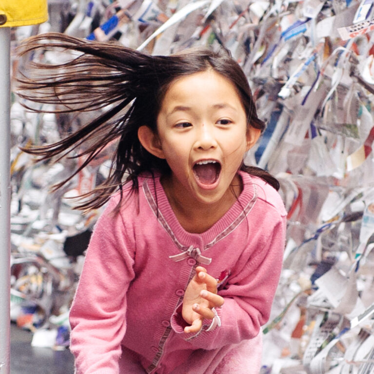 A Sticky Maze production photo. A child in a pink dress runs along a wall of sticky tape covered in hundreds of small pieces of paper. They are smiling widely and their long hair fans out behind them.