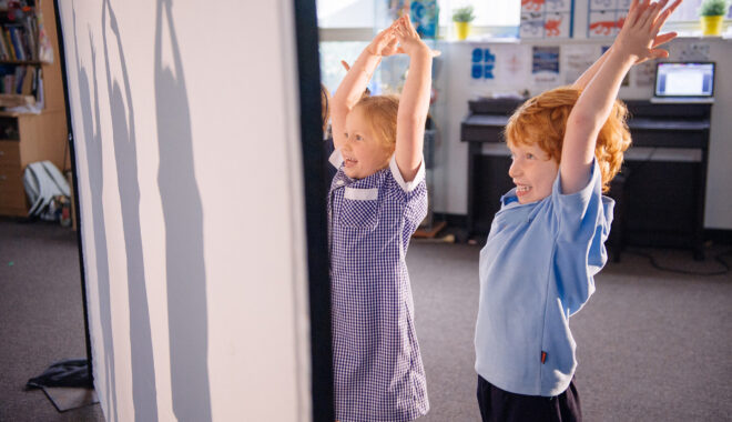 A Shadow Tricks workshop photo. Two students stand on one side of a screen in a classroom, their arms raised. Their shadows are visible on the other side of the screen.